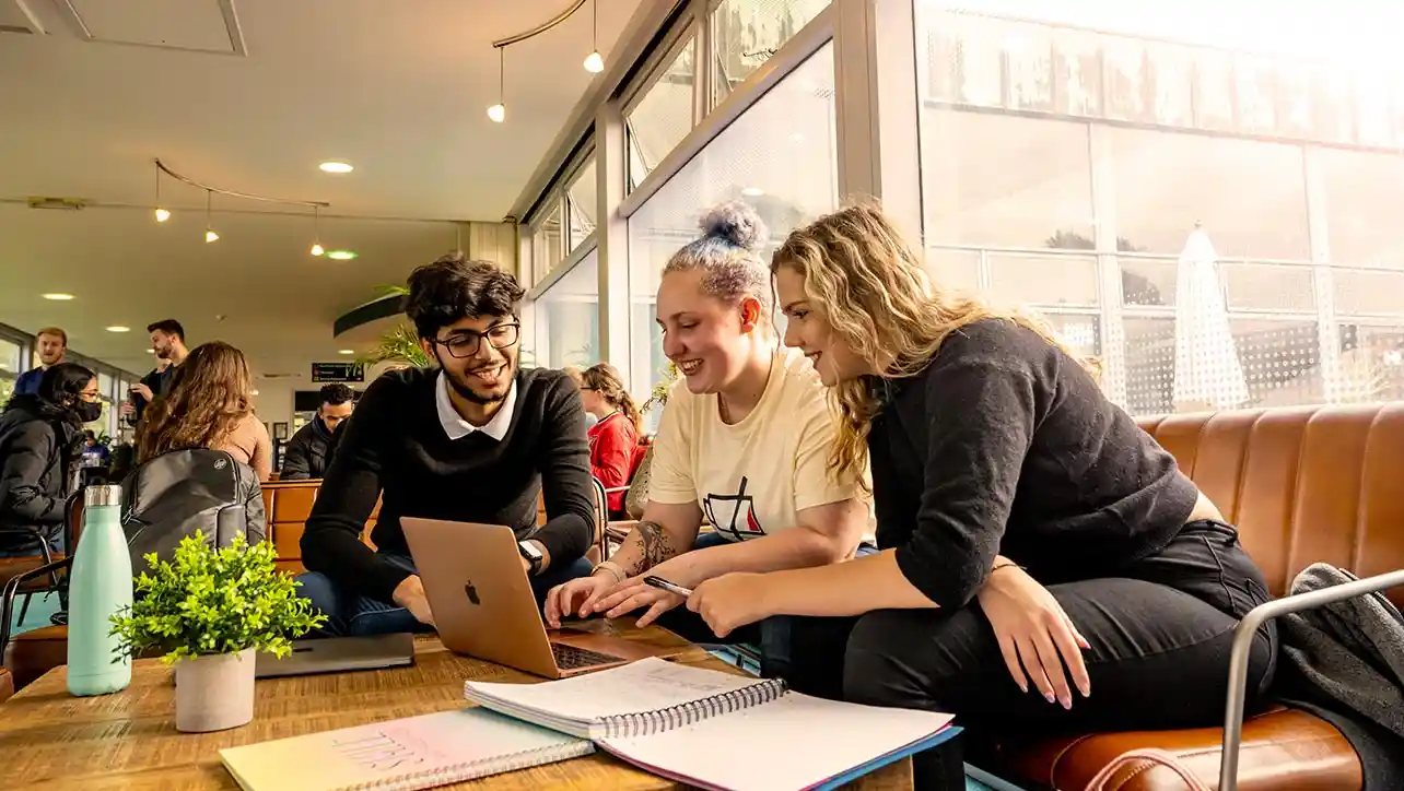 A group of three smiling students sat down, gathered around a laptop.