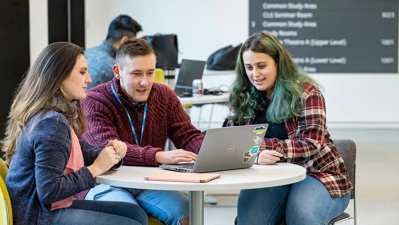 A group of three students all sat down at a round table, looking at a laptop screen.