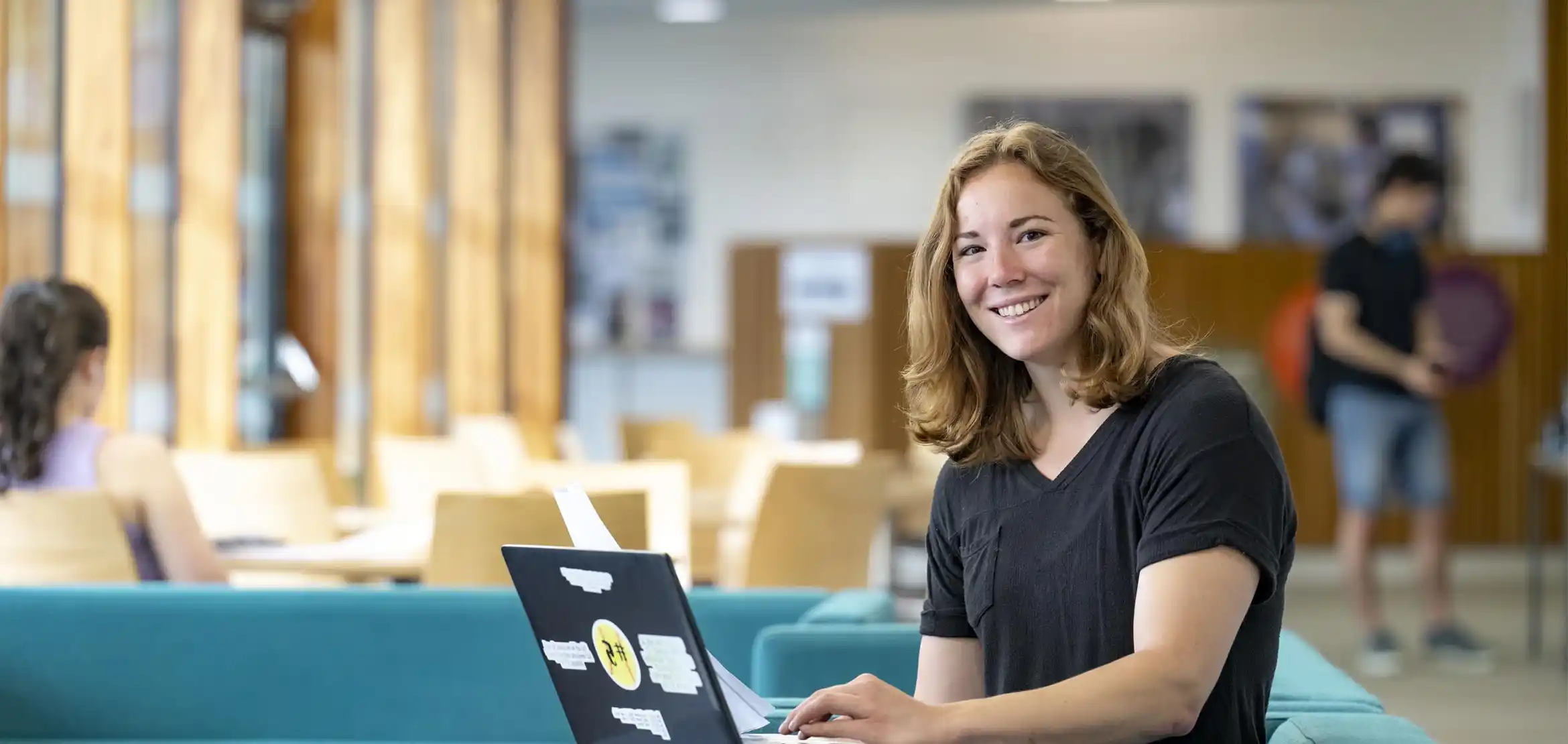 A student smiling to camera, sat down and working on a laptop.